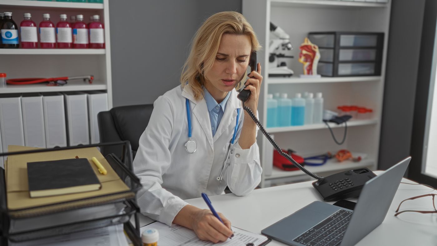 GP in a white coat on the phone at her desk, taking notes